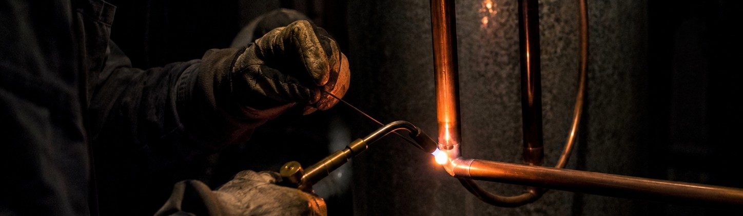 A Breakneck Mechanical technician brazing a copper line set, the torch flame lighting the copper and his gloves in a dark basement.